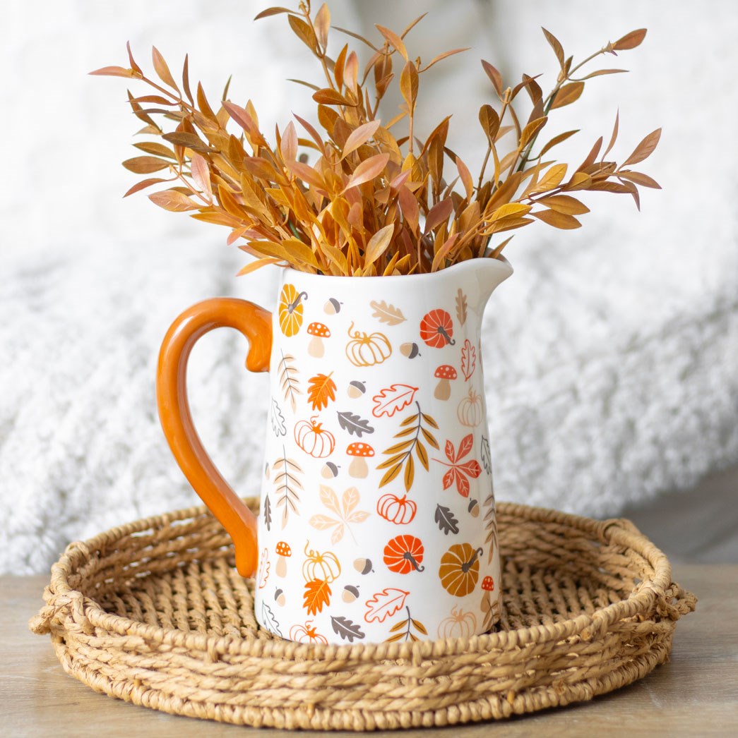 White pitcher vase with autumn-themed design and orange handle filled with dried leaves on a woven tray.