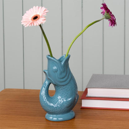 Blue fish shaped vase with flowers on a wooden surface and books in the background