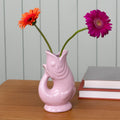 Pink ceramic fish shaped vase with two flowers on a wooden surface against a grey wall.