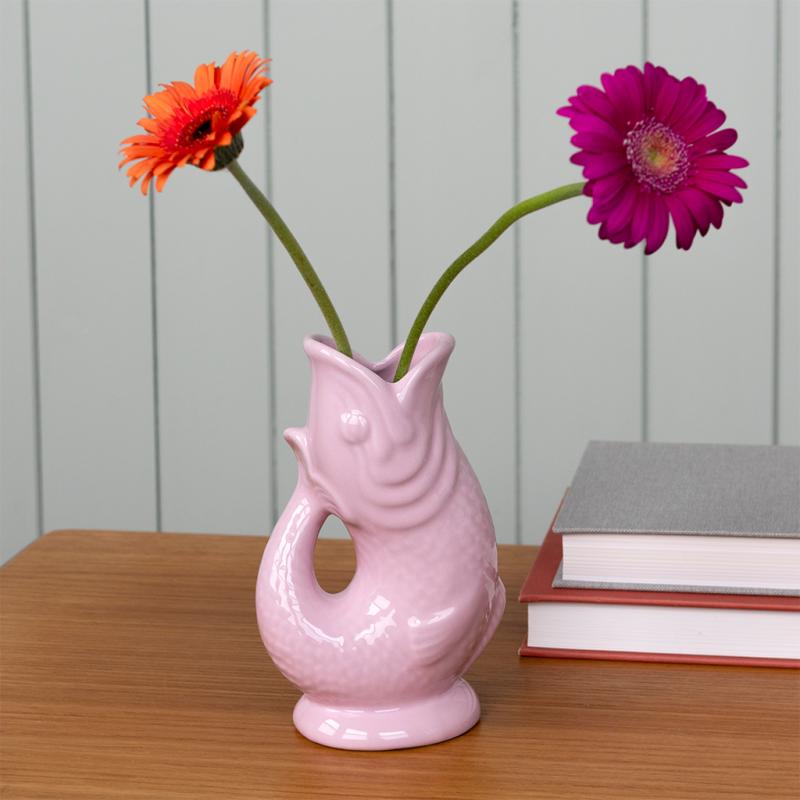 Pink ceramic fish shaped vase with two flowers on a wooden surface against a grey wall.