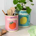 Two decorative tins labeled 'Spiced Tomatoes' and 'Moroccan Lemons' on a white surface with a plant in the background.