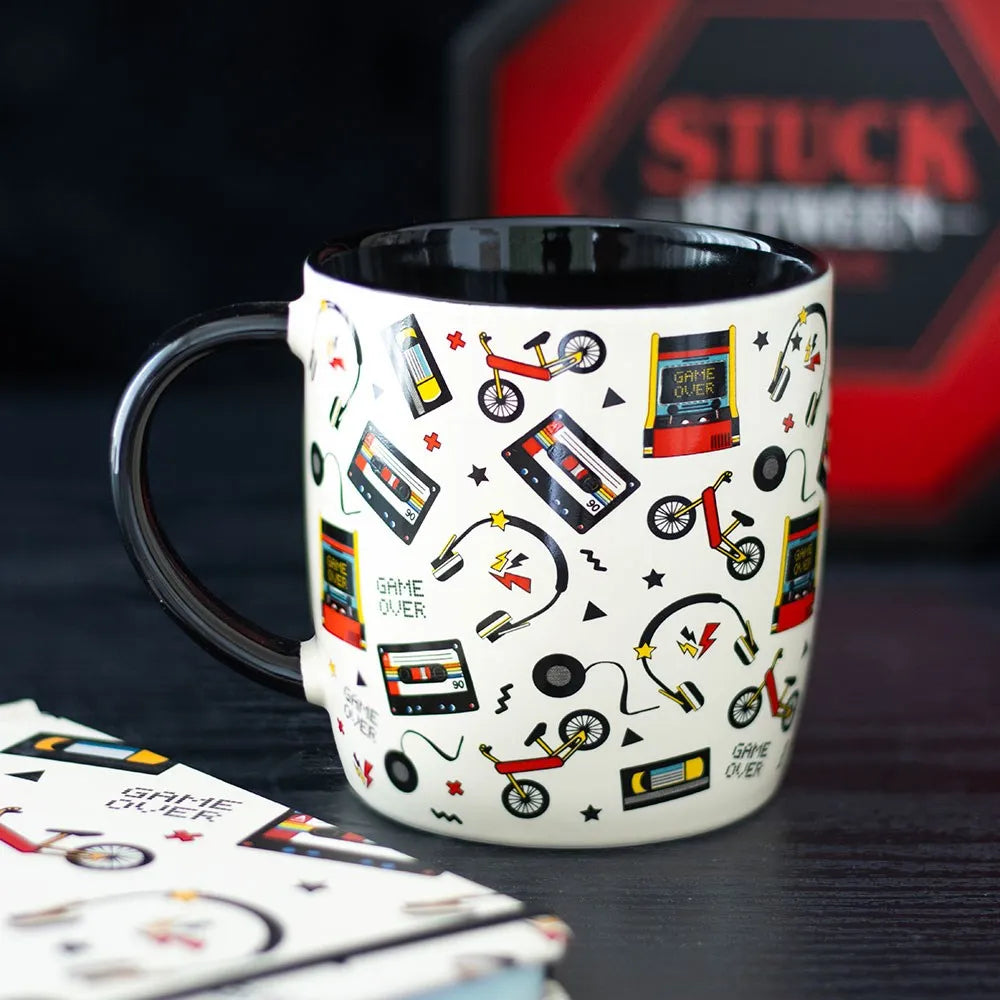 Mug with retro-themed design on a dark surface, featuring cassette tapes, game controllers, and other vintage elements.