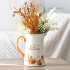 White pitcher with orange handle and 'Hello Autumn' text, filled with autumn-themed decorations on a wooden surface.