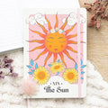 Notebook with a sun design and 'The Sun' card text on a soft surface with flowers.