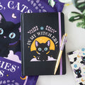 Black notebook with a cat illustration and text and a pen, surrounded by decorative items.
