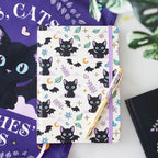 Notebook with black cat pattern on a white surface with a purple book and green leaves in the background.