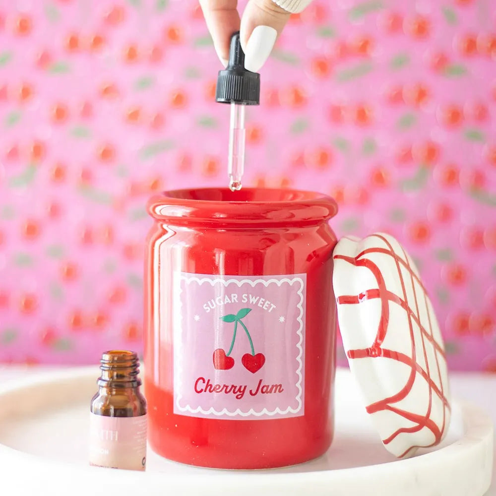 Red cherry jar oil burner labelled 'Sugar Sweet Cherry Jam' with a dropper being added to it against a pink cherry background.