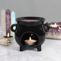 A black cauldron-shaped oil burner with cut-out star details and a lit candle inside, placed on a flat surface with crystal figures and a dimly lit background.