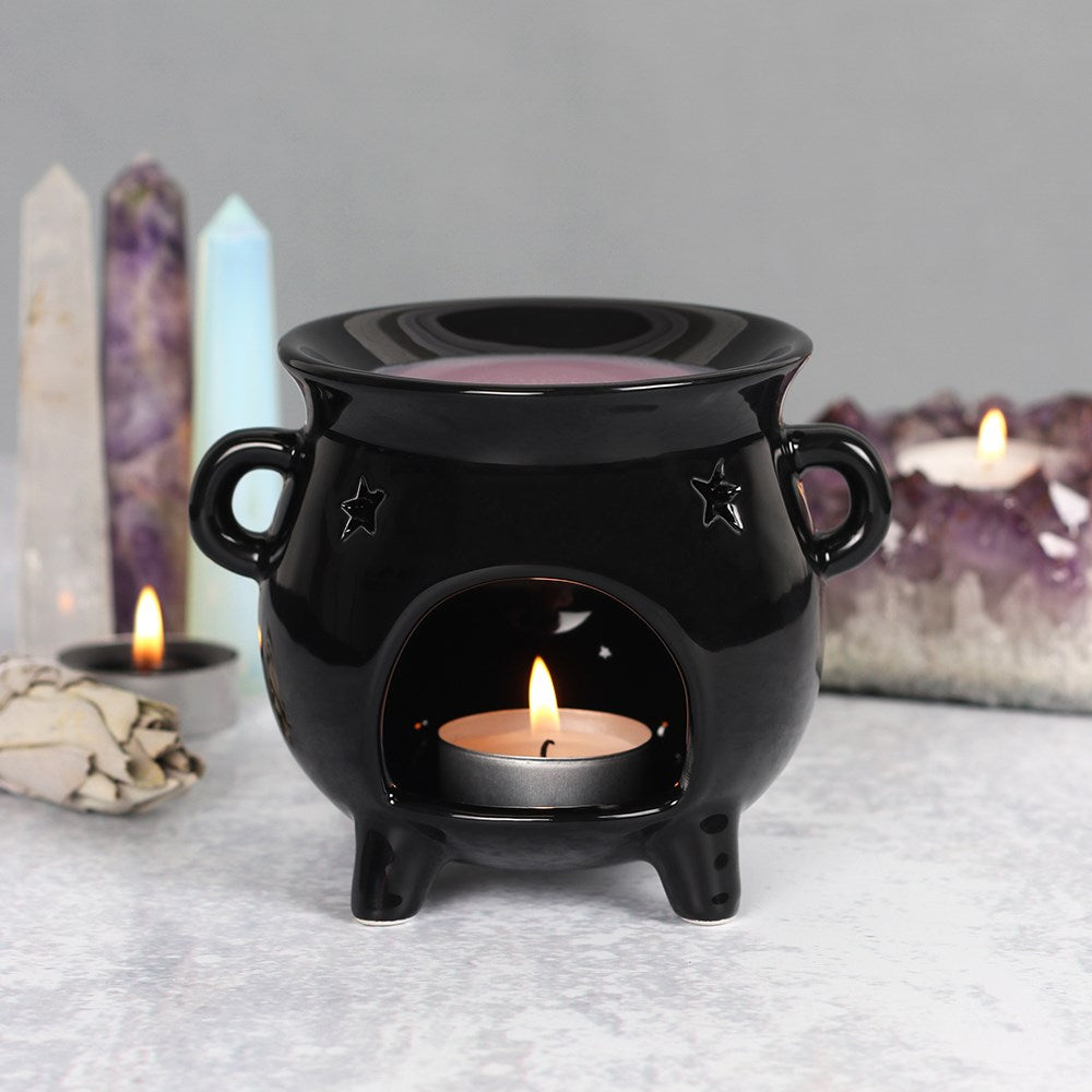 A black cauldron-shaped oil burner with cut-out star details and a lit candle inside, placed on a flat surface with crystal figures and a dimly lit background.