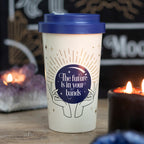 An off white bamboo eco travel mug with a navy lid and a mystical fortune teller design, placed on a wooden surface with lit candles in the background.