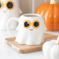 White ghost-shaped mug with sunflower sunglasses on a wooden coaster, with pumpkins in the background.