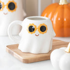 White ghost-shaped mug with sunflower sunglasses on a wooden coaster, with pumpkins in the background.