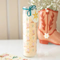Decorative Glass Tube candle with cowboy boot and cactus pattern next to a pink cowboy boot on a white surface.