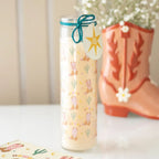Decorative Glass Tube candle with cowboy boot and cactus pattern next to a pink cowboy boot on a white surface.