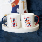 Two ceramic mugs with playing card designs on a white coaster.