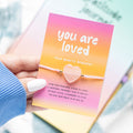 Hand holding a card with a rose quartz bracelet and 'you are loved' text.