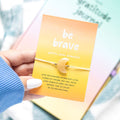 Hand holding a yellow bracelet with a 'be brave' message card.