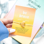 Hand holding a yellow bracelet with a 'be brave' message card.
