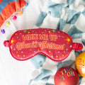 Red sleep mask with 'Wake Me Up When It's Christmas' text on a patterned blanket.