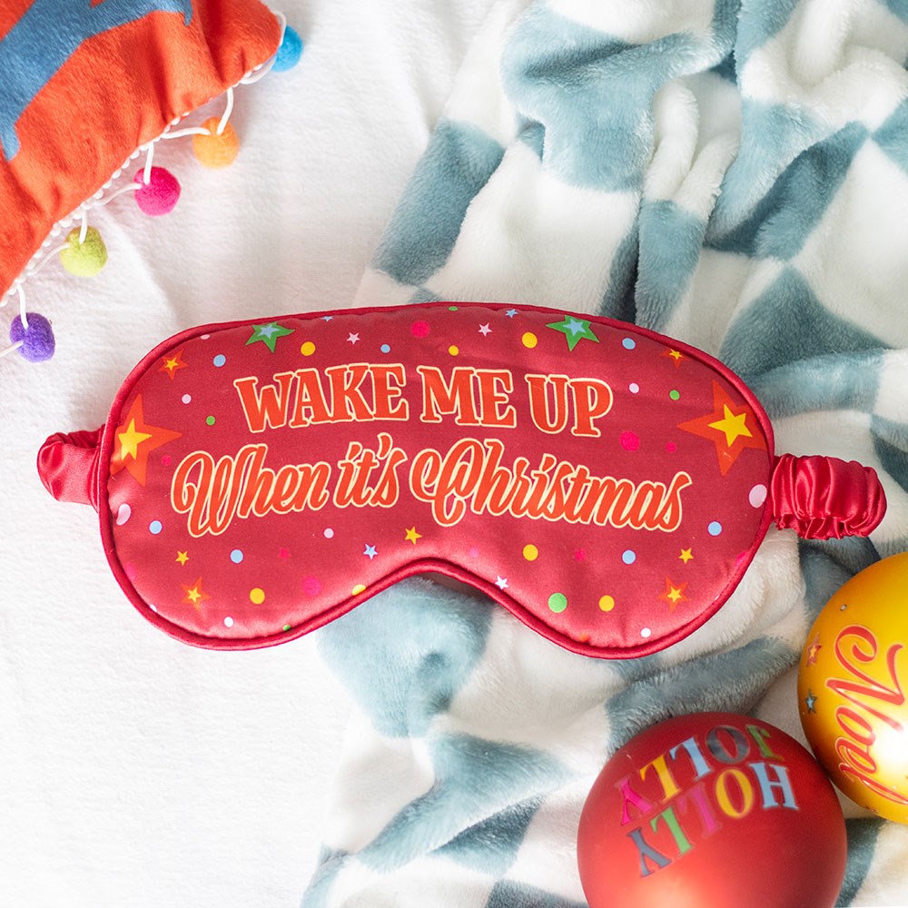 Red sleep mask with 'Wake Me Up When It's Christmas' text on a patterned blanket.
