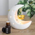 white ceramic Moon-shaped oil burner candle holder with a small bottle of essential oil on a wooden surface.