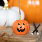Small orange pumpkin with a face incense stick holder on a wooden surface with blurred pumpkins in the background.