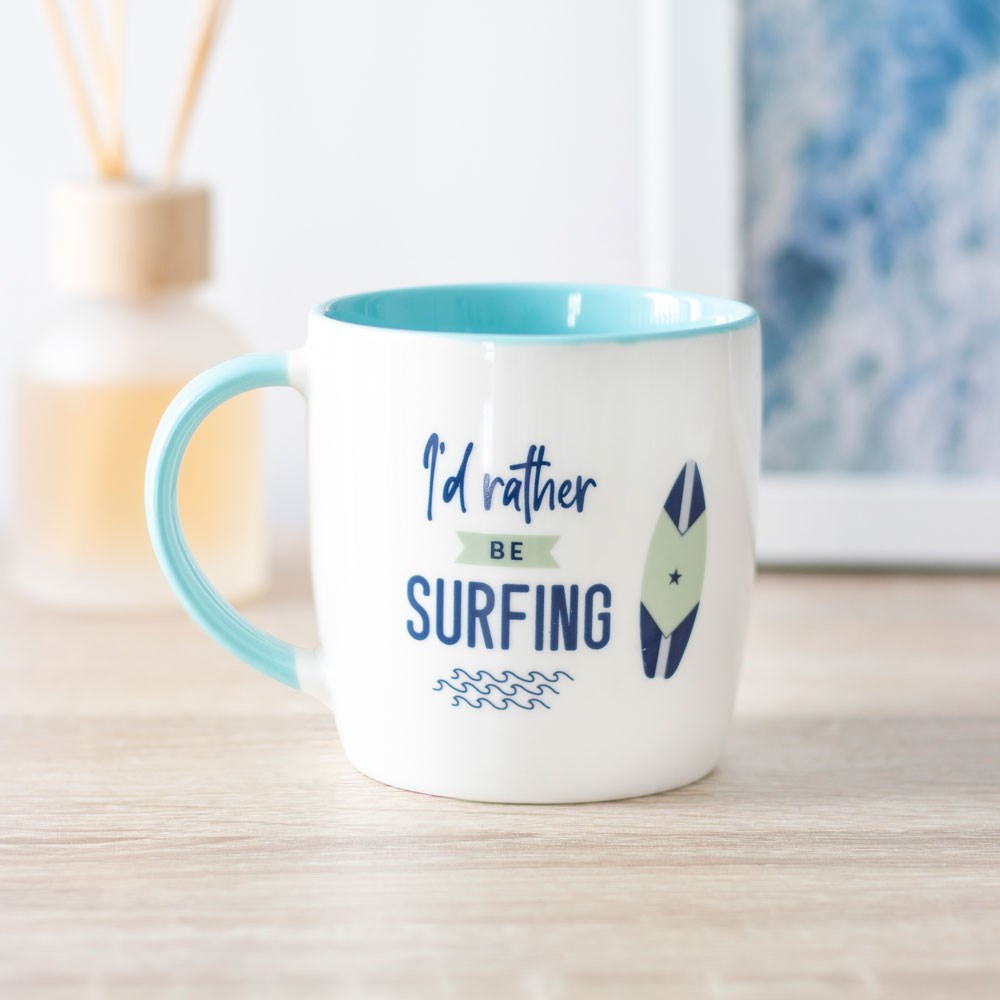 A white ceramic mug with a teal handle and inside, featuring the text 'I'd Rather Be Surfing' and illustrations of a surfboard and waves.