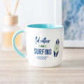 A white ceramic mug with a teal handle and inside, featuring the text 'I'd Rather Be Surfing' and illustrations of a surfboard and waves.