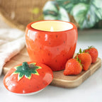 Ceramic strawberry-shaped candle with a lit wick on a wooden board with strawberries.