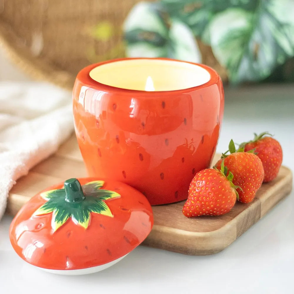 Ceramic strawberry-shaped candle with a lit wick on a wooden board with strawberries.