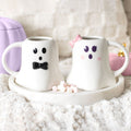 Two ceramic mugs shaped like a ghost with a bow tie and hair bow on a white surface.