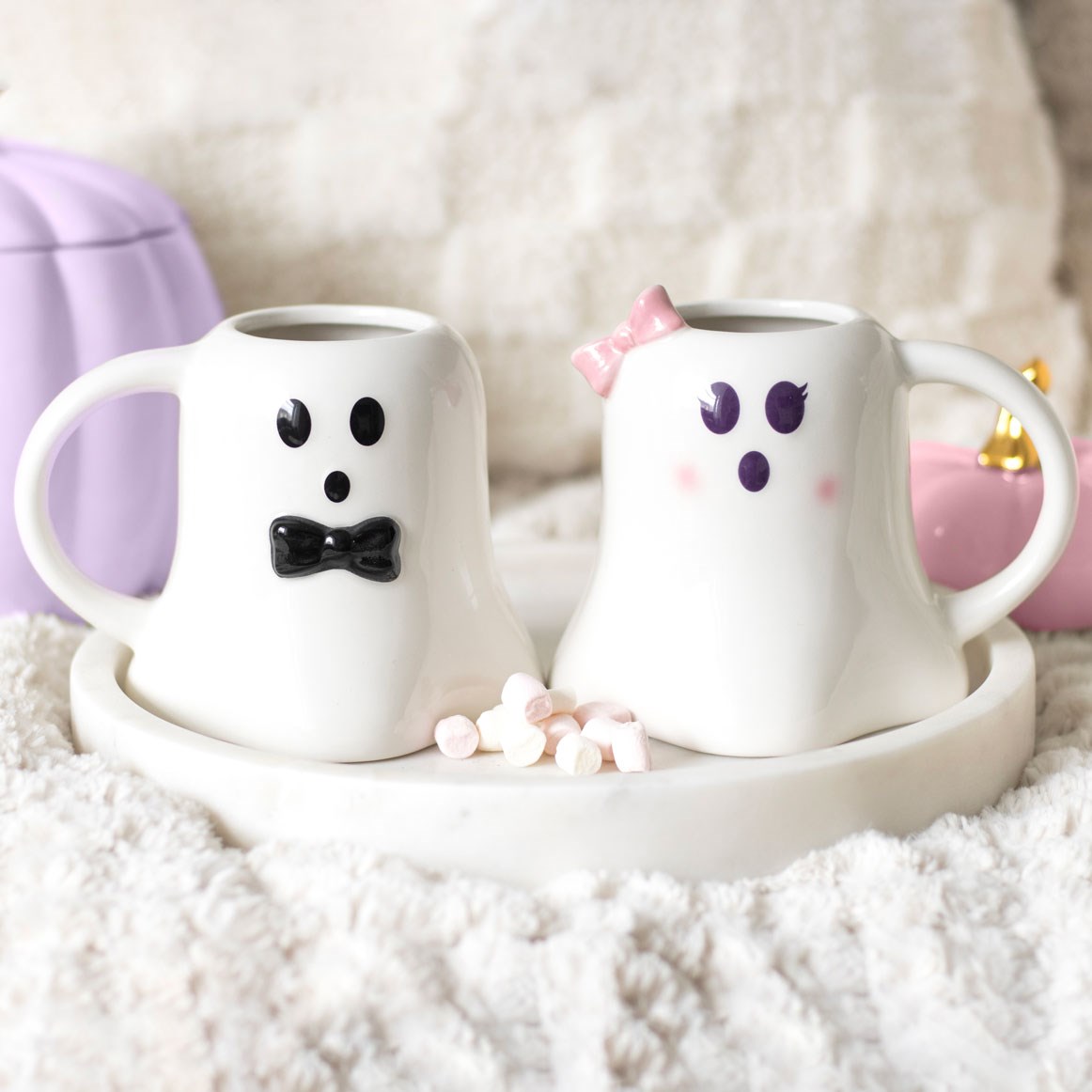Two ceramic mugs shaped like a ghost with a bow tie and hair bow on a white surface.