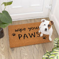 Dog sitting on a coir doormat with 'wipe your PAWS' text in a home setting.