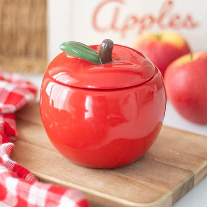Red apple-shaped candle jar with a lid on a wooden cutting board with apples in the background.