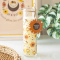 Glass tube candle with floral  and bee design and 'Wildflower Scent' label on a surface with a plant in the background