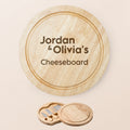 Personalised round wooden cheeseboard set with engraved text reading 'Jordan & Olivia's Cheeseboard' on a light beige background, shown with the small knife and cheese spreader accessories below.