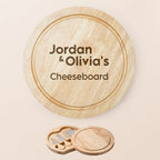 Personalised round wooden cheeseboard set with engraved text reading 'Jordan & Olivia's Cheeseboard' on a light beige background, shown with the small knife and cheese spreader accessories below.