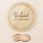 Personalised round wooden cheese board set with engraved name and quote 'Still maturing since...', shown on a light beige background with the matching wooden cheese knife box below.