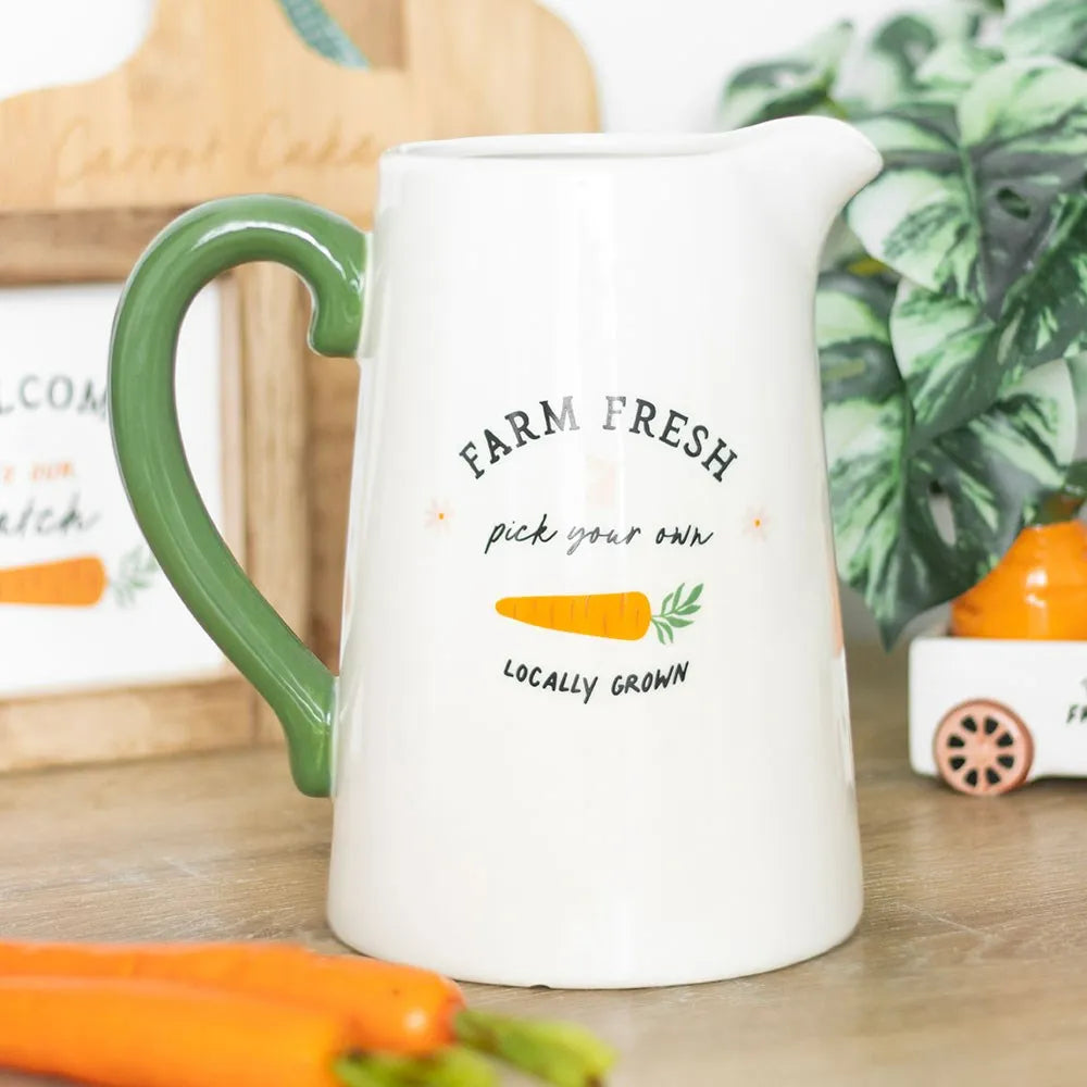 White pitcher style vase with green handle and 'Farm Fresh' text on a wooden surface with carrots and plants.