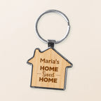 A house-shaped wooden keychain with a silver metal split ring, featuring the engraved text 'Maria's HOME Sweet HOME' in brown lettering on a light wood grain background.
