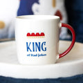 White mug with red handle featuring 'King of Dad Jokes' text on a blurred background