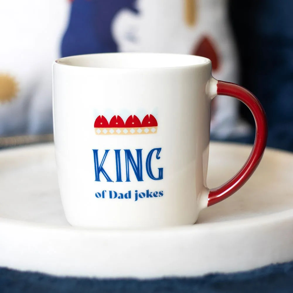 White mug with red handle featuring 'King of Dad Jokes' text on a blurred background