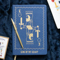 Blue notebook with King of Hearts design and gold pen on a dark blue textured background