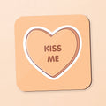 A square coaster with rounded corners featuring a candy heart design. The coaster has a peach-beige background, a white border, and a brown heart outline in the center containing the words 'KISS ME' in brown.