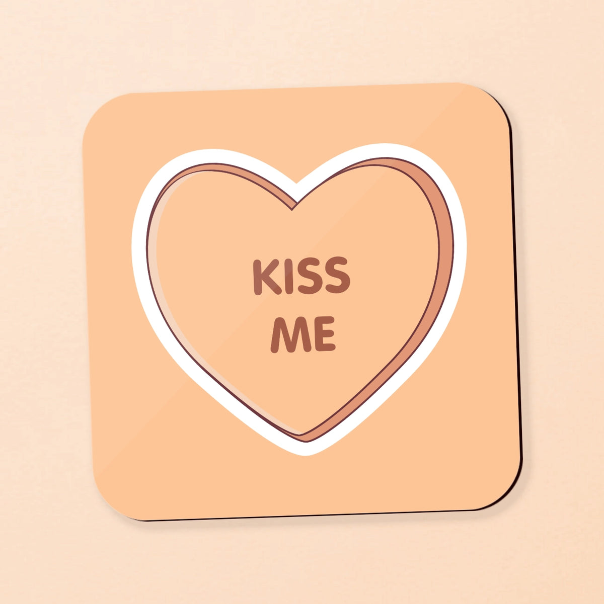 A square coaster with rounded corners featuring a candy heart design. The coaster has a peach-beige background, a white border, and a brown heart outline in the center containing the words 'KISS ME' in brown.