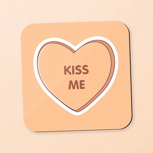 A square coaster with rounded corners featuring a candy heart design. The coaster has a peach-beige background, a white border, and a brown heart outline in the center containing the words 'KISS ME' in brown.