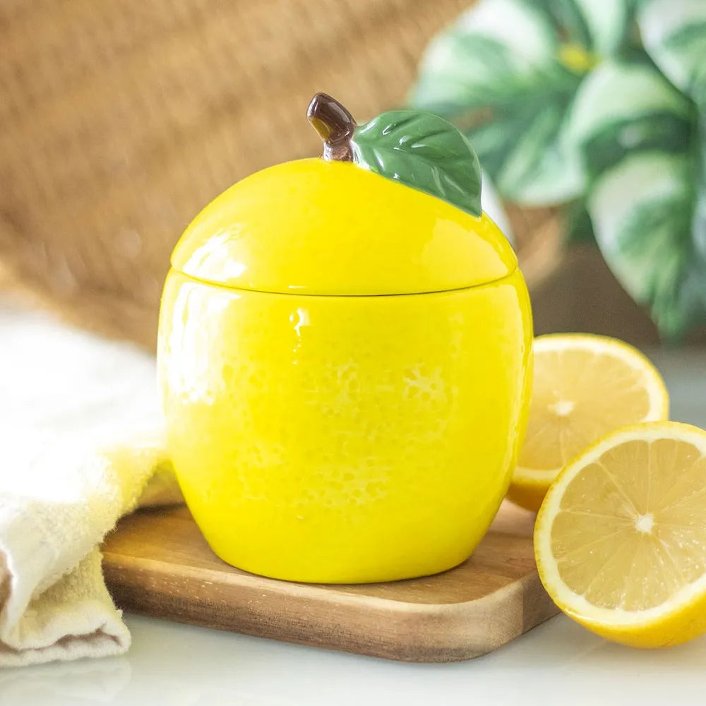 Yellow lemon-shaped container candle with a lid on a wooden board with lemons in the background.