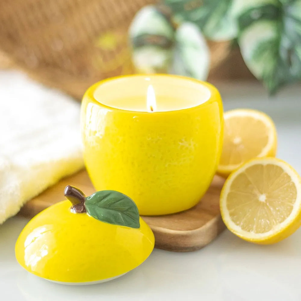 Yellow ceramic candle with a lemon design on a wooden coaster, surrounded by lemons and greenery.