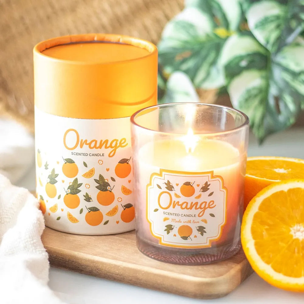 Orange scented candle in a glass jar and cylindrical packaging container with oranges and leaves in the background.