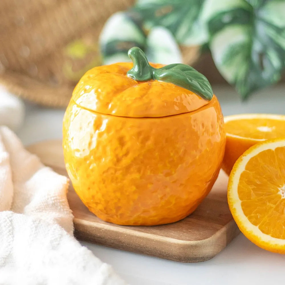 Orange ceramic candle container with a lid resembling an orange, placed on a wooden board with a white cloth and green leaves in the background.
